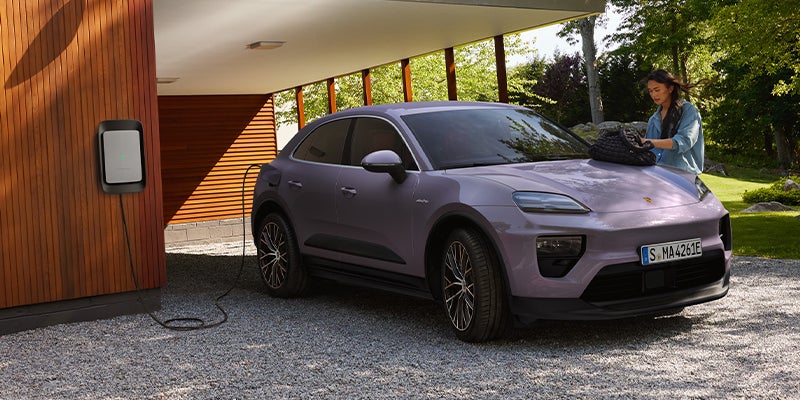 A dreams provence 2025 Porsche All- Electric Macan 4 parked and charging in a residential driveway while a woman sifts through her purse that is setting on the hood of the vehicle on a sunny day